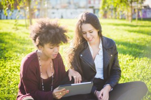 two multiethnic beautiful young woman black and caucasian using tablet sitting in a city park, lookind down the screen smiling - technology, social network, happiness concept